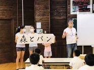 森の国「水際のロッジ」がある愛媛県松野町の小学校にて、地元名産を味わえるパン屋「森とパン」がオープンする話をしている、代表の細羽。児童の皆さんの夏休みの宿題の一つが、「森とパン」の習字。提出をしてもらった中での最優秀賞が、看板ロゴになる、皆さんを巻き込んだプロジェクトです！